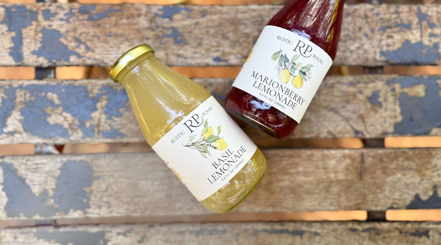Two bottles of Rustic Pours lemonade sitting on a rustic park bench; one bottle reads Basil Lemonade, the other reads Marionberry Lemonade