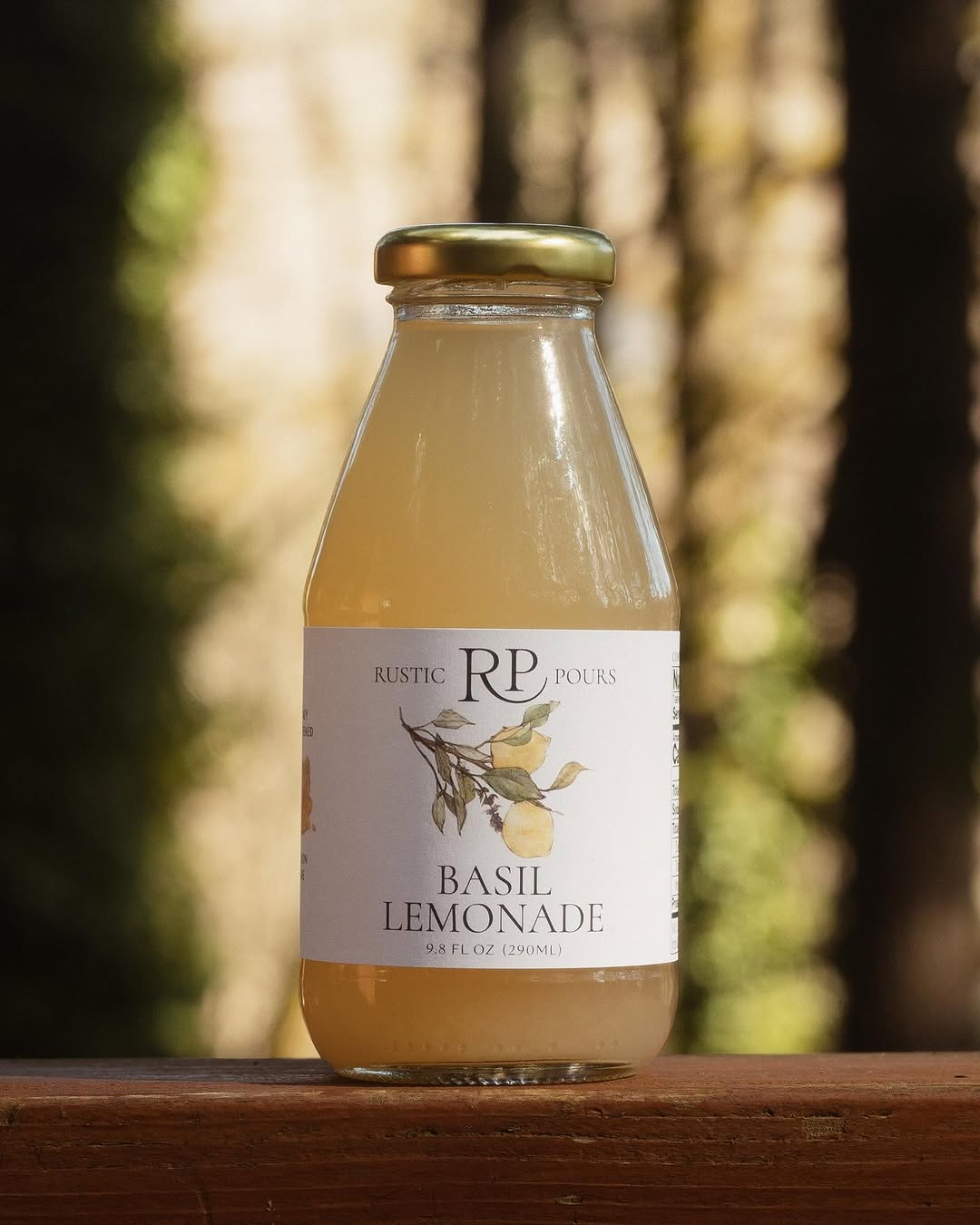 A glass bottle of Rustic Pours Basil Lemonade, with gold cap and watercolor illustration on label, sitting on a wood surface with tall trees blurred in the background.