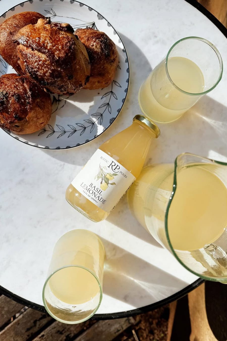 A white marble table seen from above, with a plate of croissants and Rustic Pour Basil Lemonade in a bottle and served in two glasses.