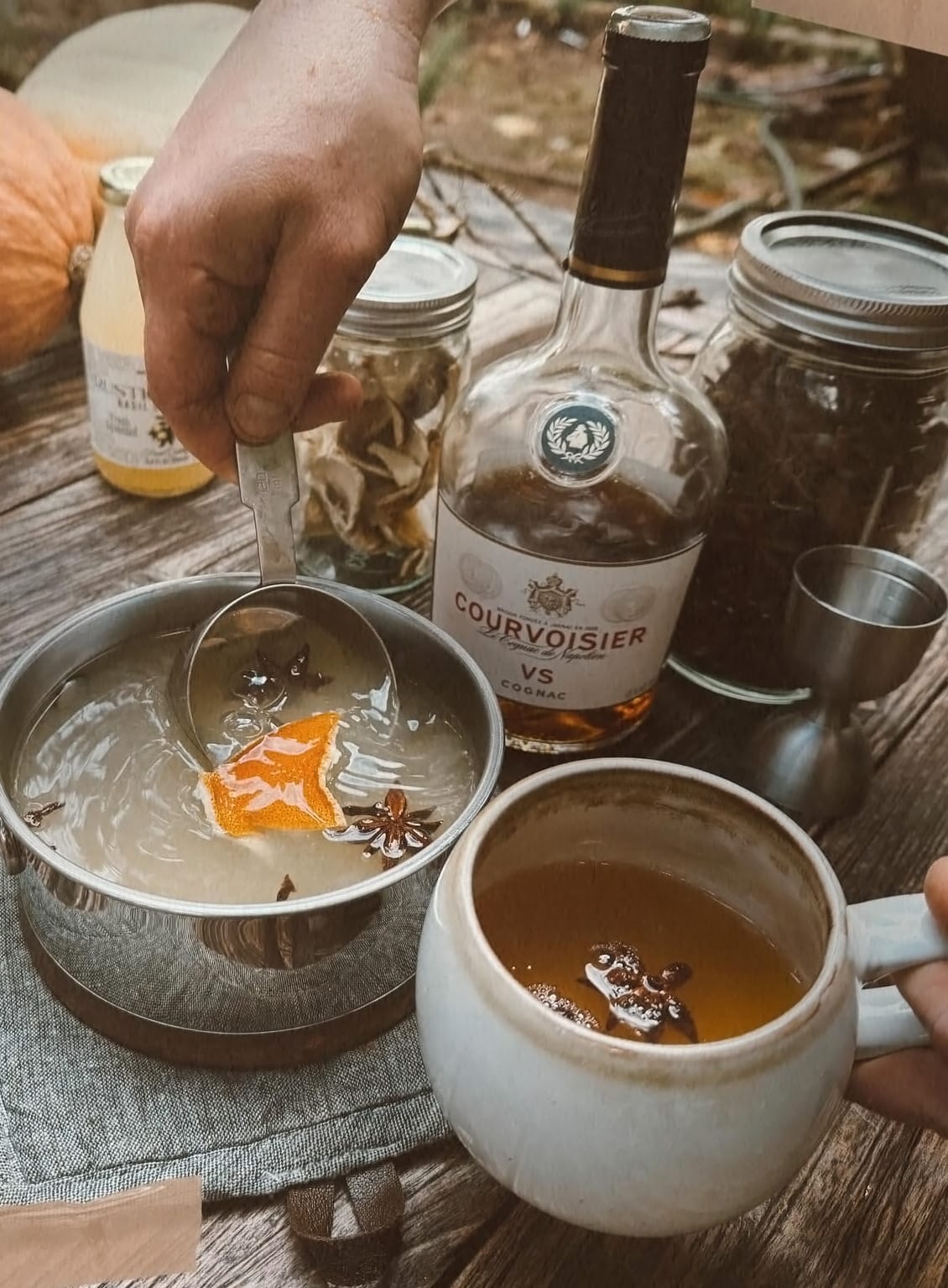 A man's hand scooping a serving of basil lemonade hot toddy into a mug, with a bottle of Courvoisier and fall gourds on a wooden outdoor table.