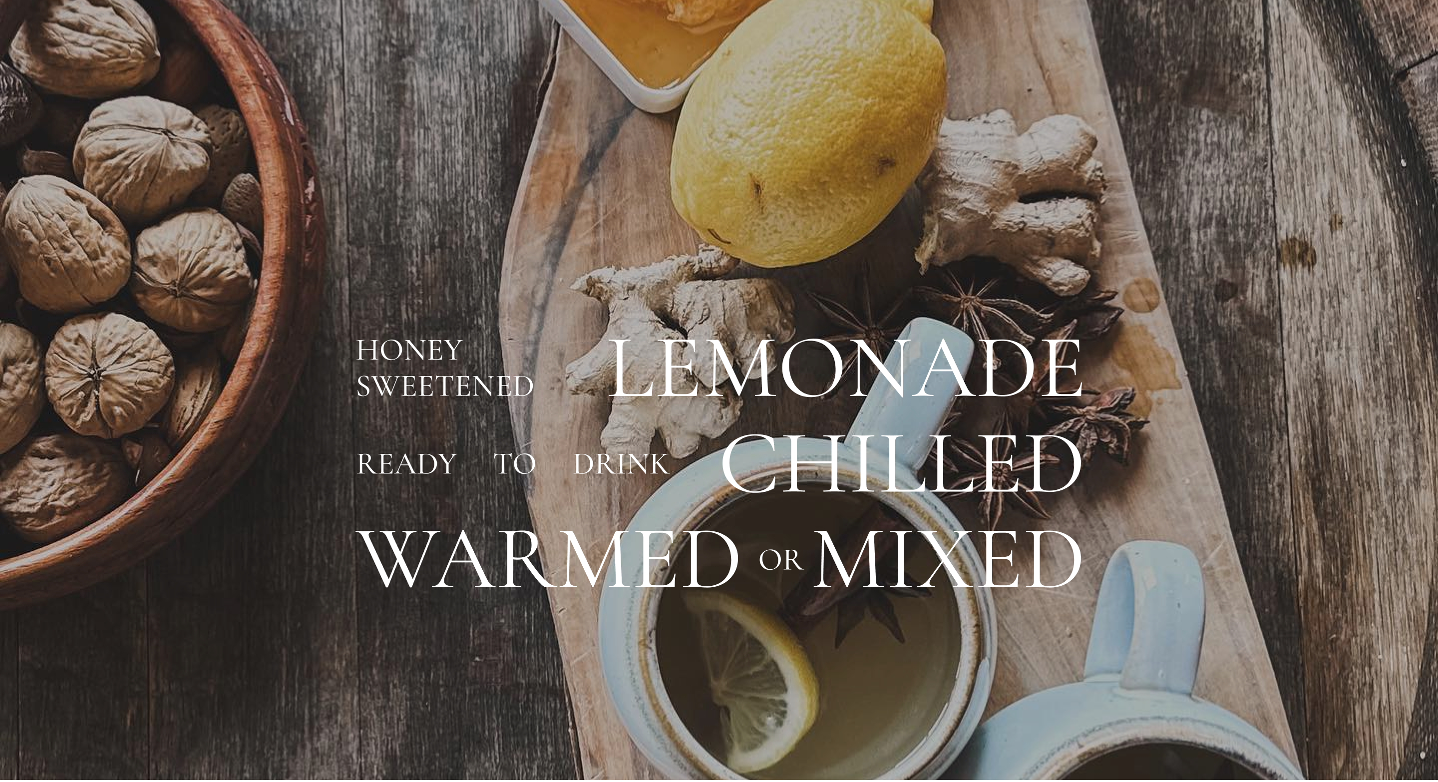 A rustic wood board holding a dish of honeycomb, a lemon, pieces of ginger and anise seed, and two mugs of hot lemonade. Text overlay reads Honey sweetened lemonade, ready to drink chilled, warmed, or mixed.