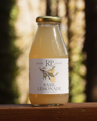 A glass bottle of Rustic Pours Basil Lemonade, with gold cap and watercolor illustration on label, sitting on a wood surface with tall trees blurred in the background.