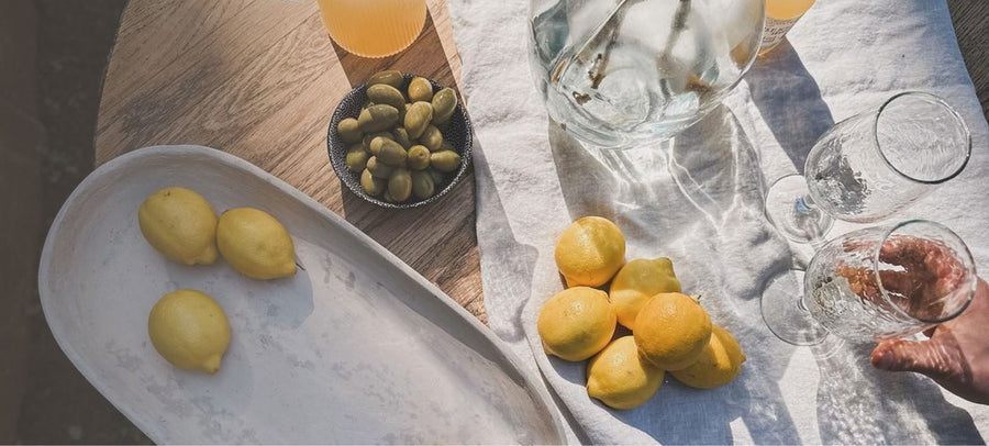 A tablescape from above featuring a bowl of lemons, glassware, and bowl of olives on a white linen cloth.