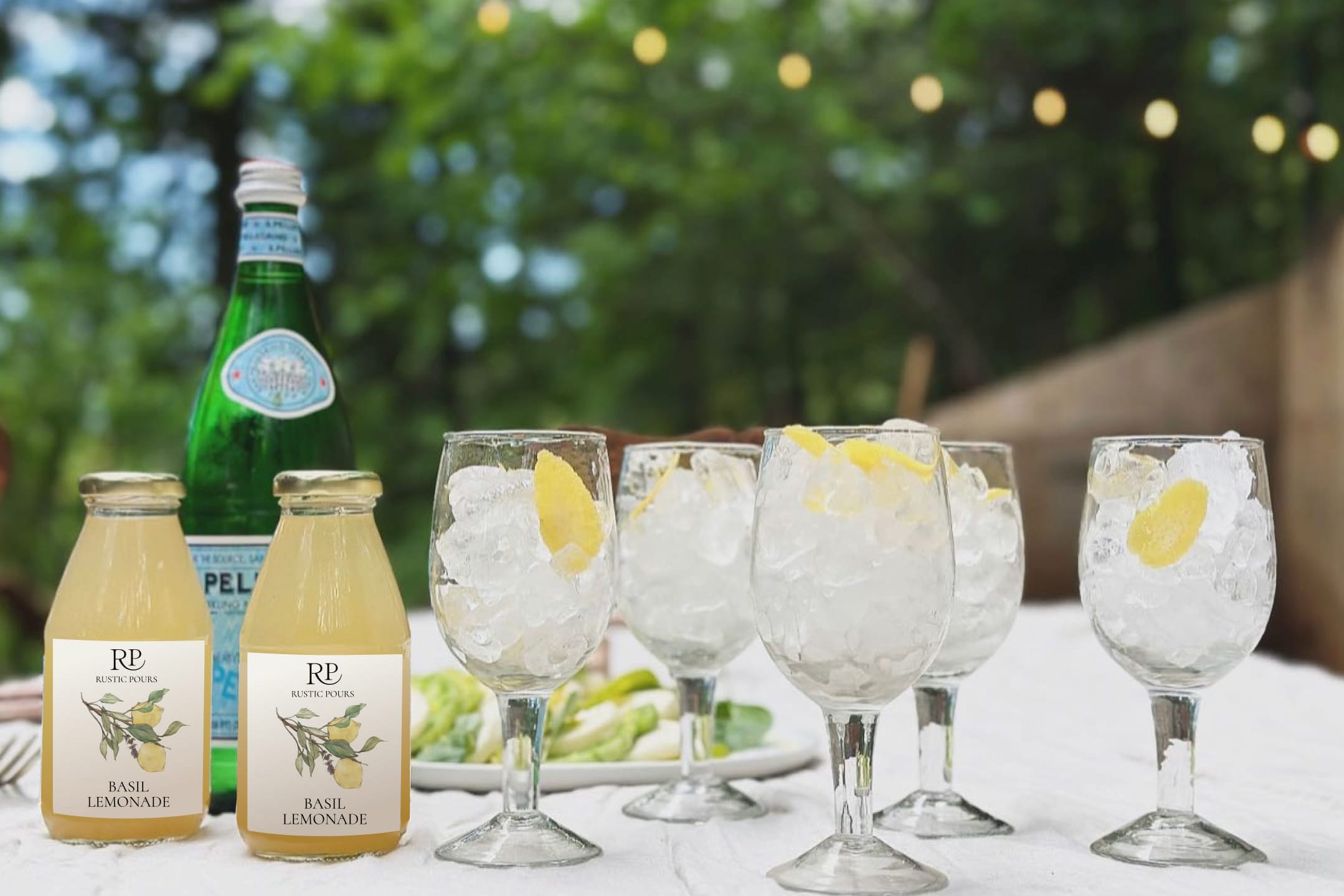 Two bottles of Rustic Pours basil lemonade on a table with sparkling water and a row of stemmed glasses filled with ice; trees and twinkle lights in the background.